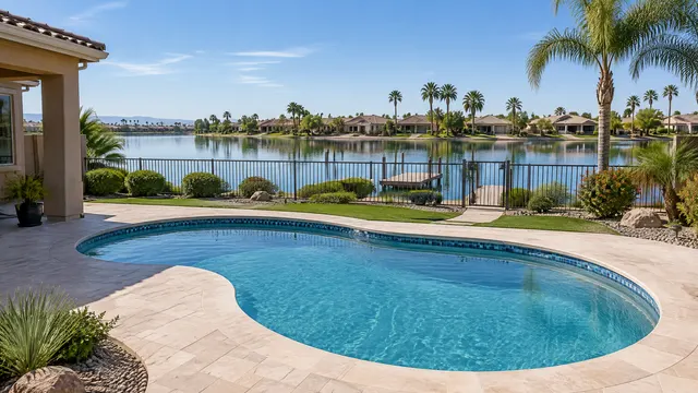 Lakefront backyard in a Gilbert, Arizona lake community: a modest kidney-shaped pool with new tile and coping, a low wrought-iron fence and gate between the yard and a calm lake with a small dock and palm trees beyond