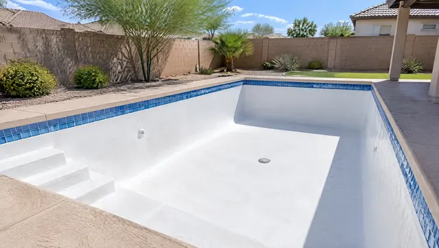 Drained rectangular backyard pool with a freshly acid-washed bright white plaster interior, clean blue waterline tile, dry steps and a main drain visible, sunny Gilbert, Arizona yard with a block wall