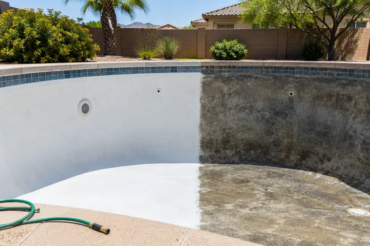 Half of a drained pool wall freshly acid washed bright white beside the untreated dark half, clean dividing line, garden hose on the deck