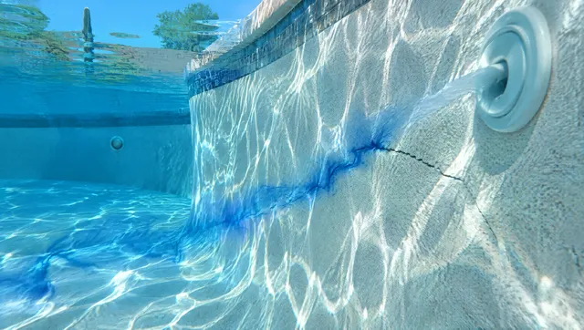 Close view of a backyard pool wall under clear water with a small dye stream being drawn toward a crack near a return jet, sunlight ripples on a light plaster finish, Gilbert, Arizona backyard