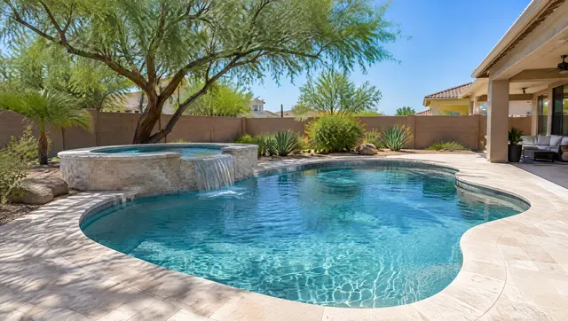 Renovated free-form backyard pool in a Gilbert, Arizona yard: new aqua pebble finish, a new raised spa wall with a sheer water spill into the pool, light travertine-look coping and deck, mature mesquite tree shading part of the patio, clear blue sky